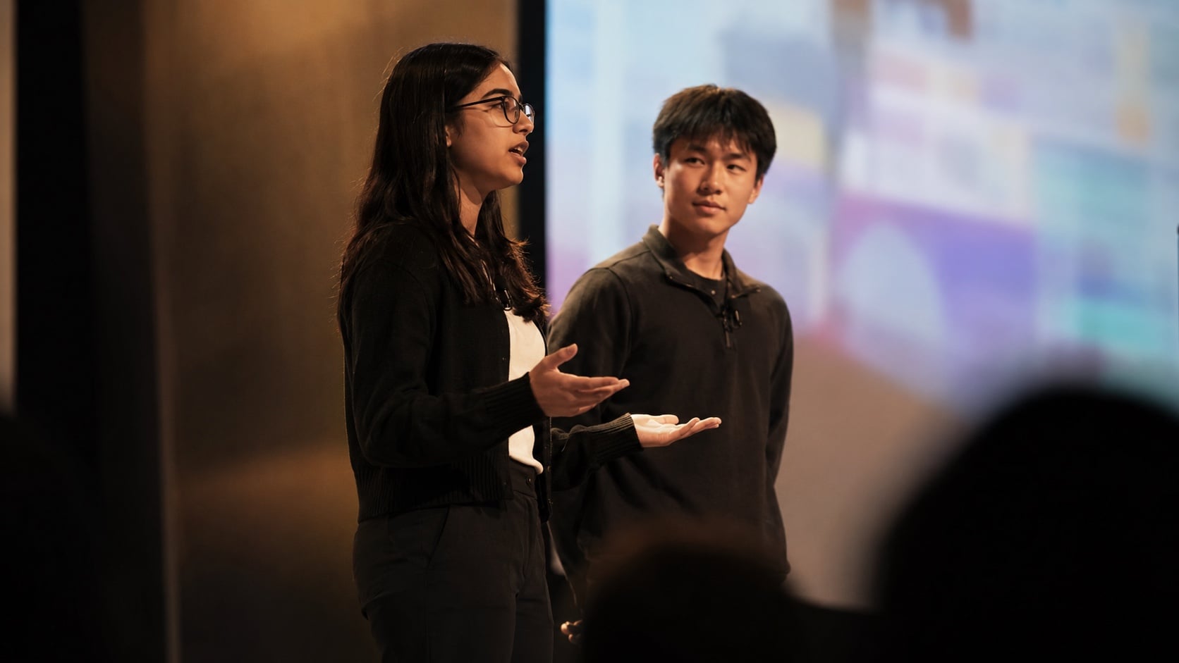 Eight high school students presenting AI projects on large screens to an audience of parents and faculty in a modern auditorium under warm stage lighting
